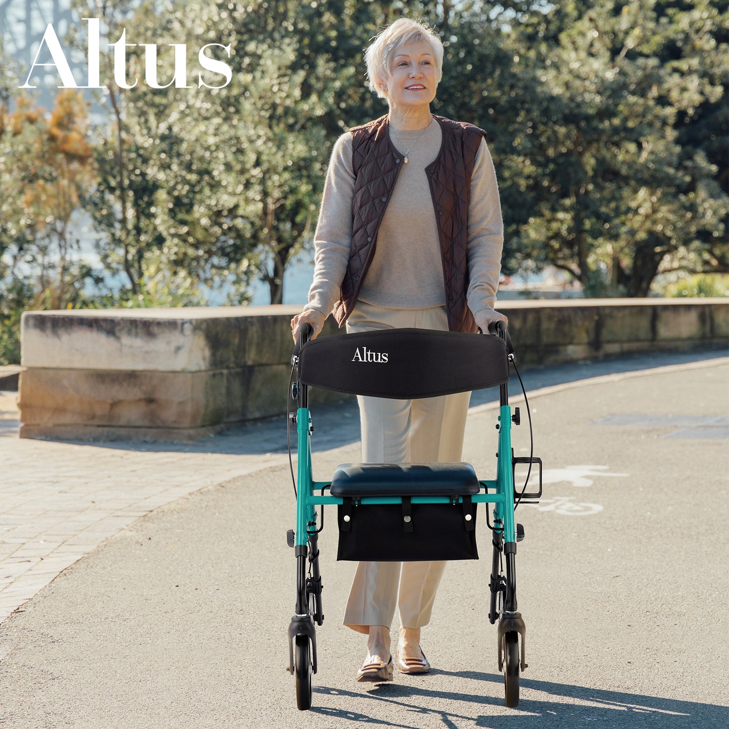 An elderly woman is walking outdoors using an Altus walker with a seat. The walker features a red frame, a black seat, and a storage bag, blending seamlessly with the natural environment.