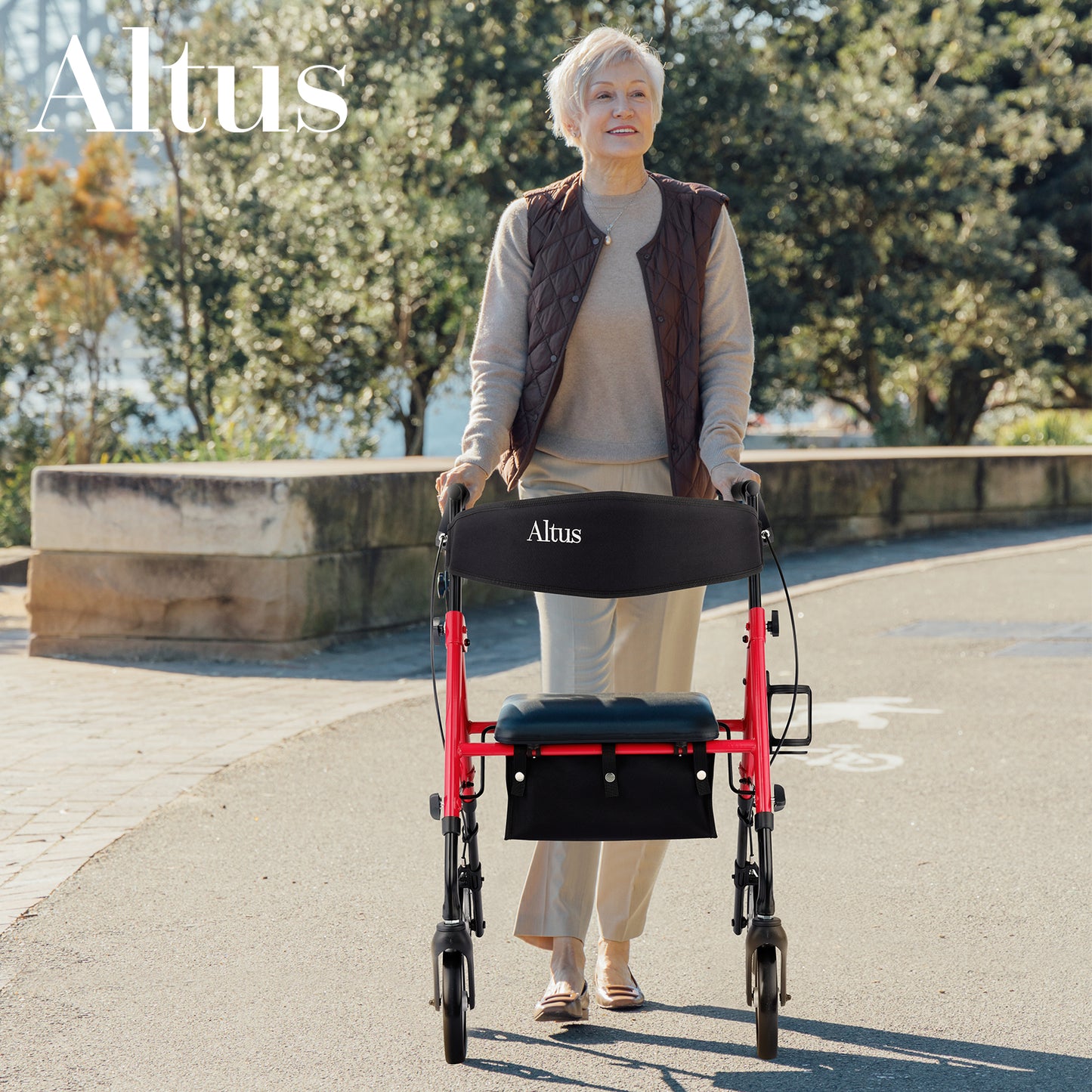 An elderly woman is walking outdoors using an Altus walker with a seat. The walker features a red frame, a black seat, and a storage bag, blending seamlessly with the natural environment.
