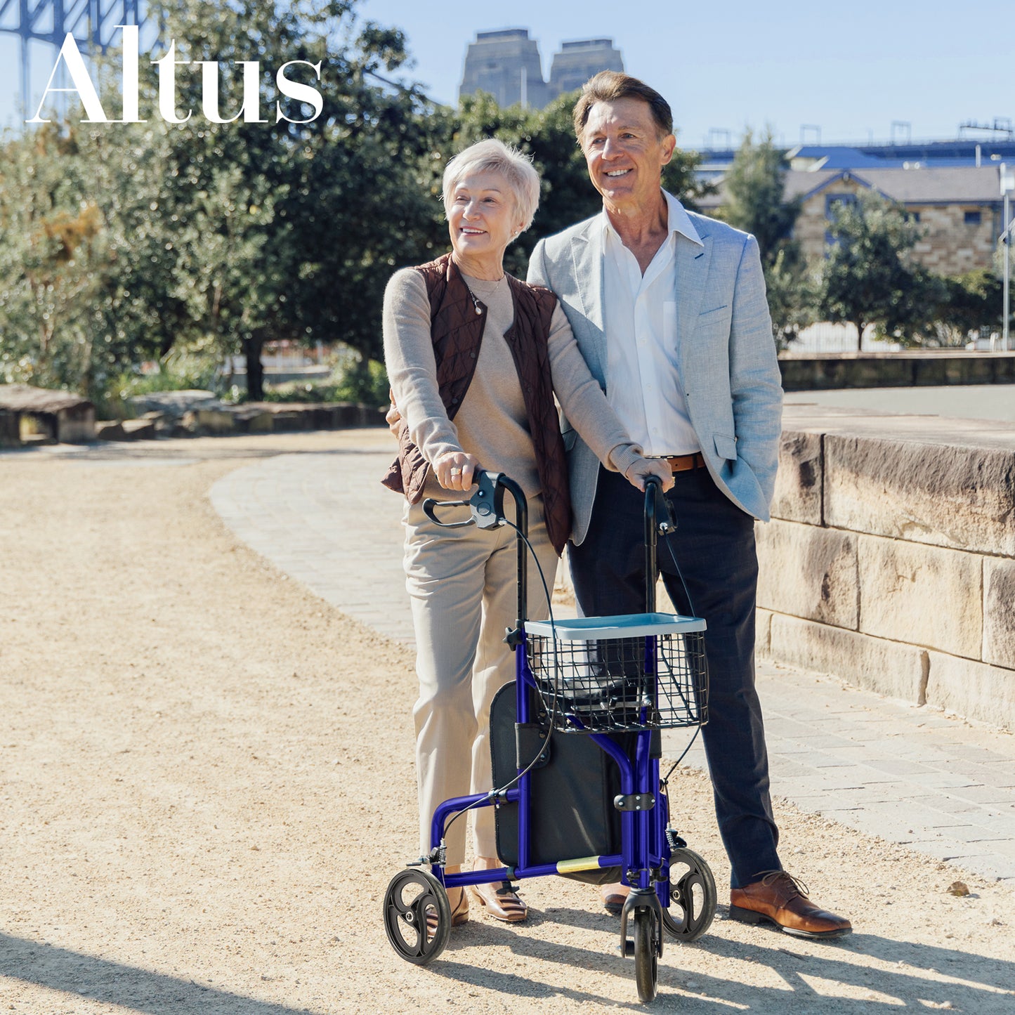Man pushing a woman in a walker along a path with trees and buildings in the background.
