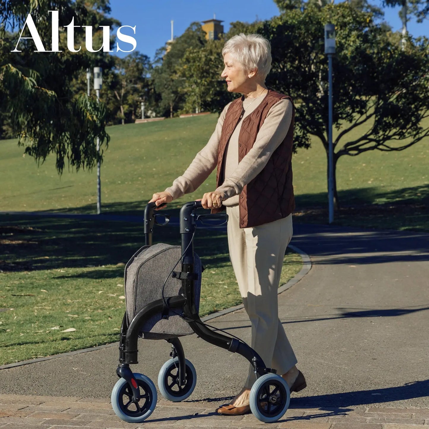 A senior woman using the Altus Folding 3 Wheel Walker Tri-Rollator in a park. This rollator includes an 8-inch wheel for smooth navigation and a durable cloth storage bag for convenience, ideal for active seniors on the go