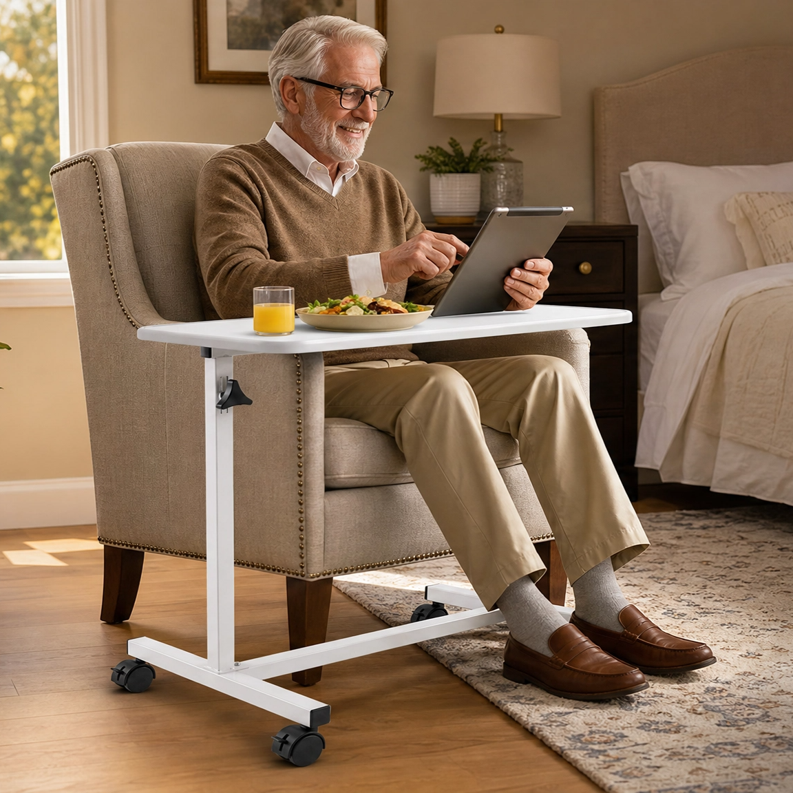 A man sits in a beige upholstered armchair in a bedroom setting, using a white adjustable lap desk with a tablet and a glass of orange juice. The lap desk has casters and is shown in a real home environment.