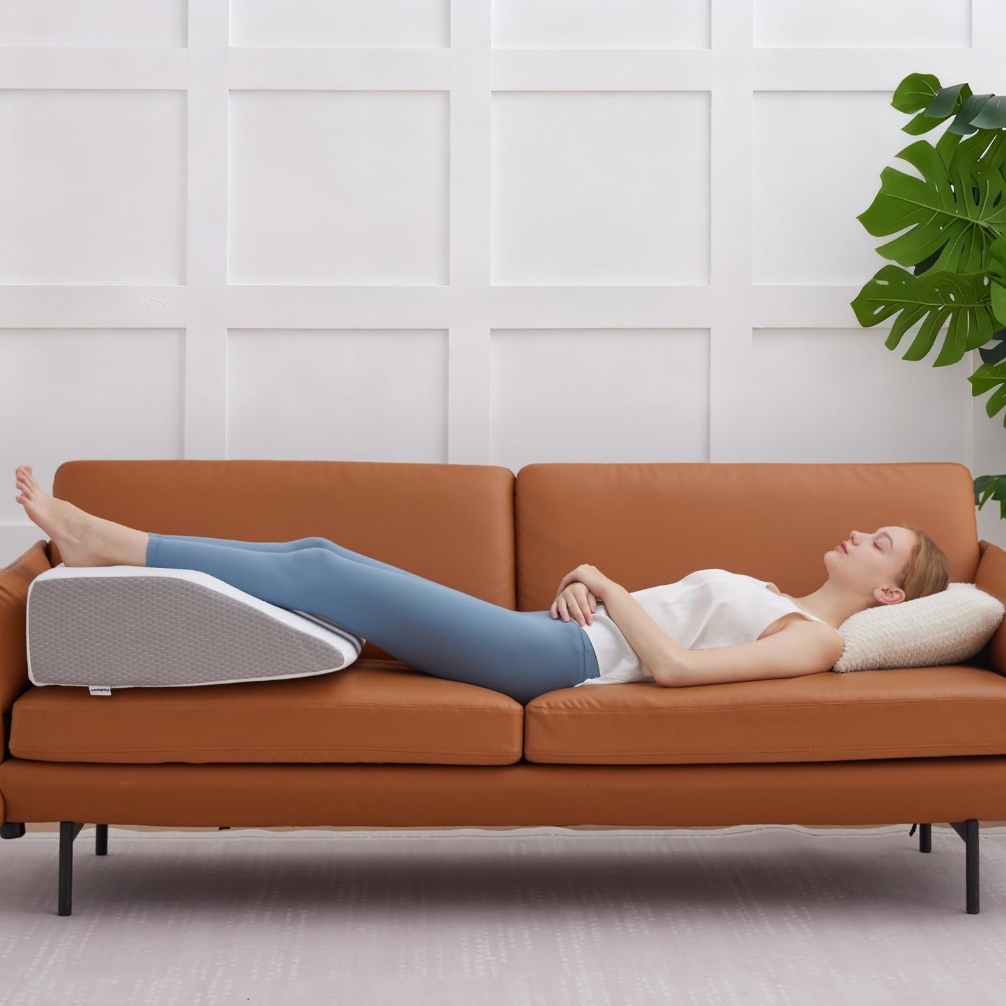 Woman lying on a brown couch with a wedge pillow under her legs, in a room with a white paneled wall and a plant.