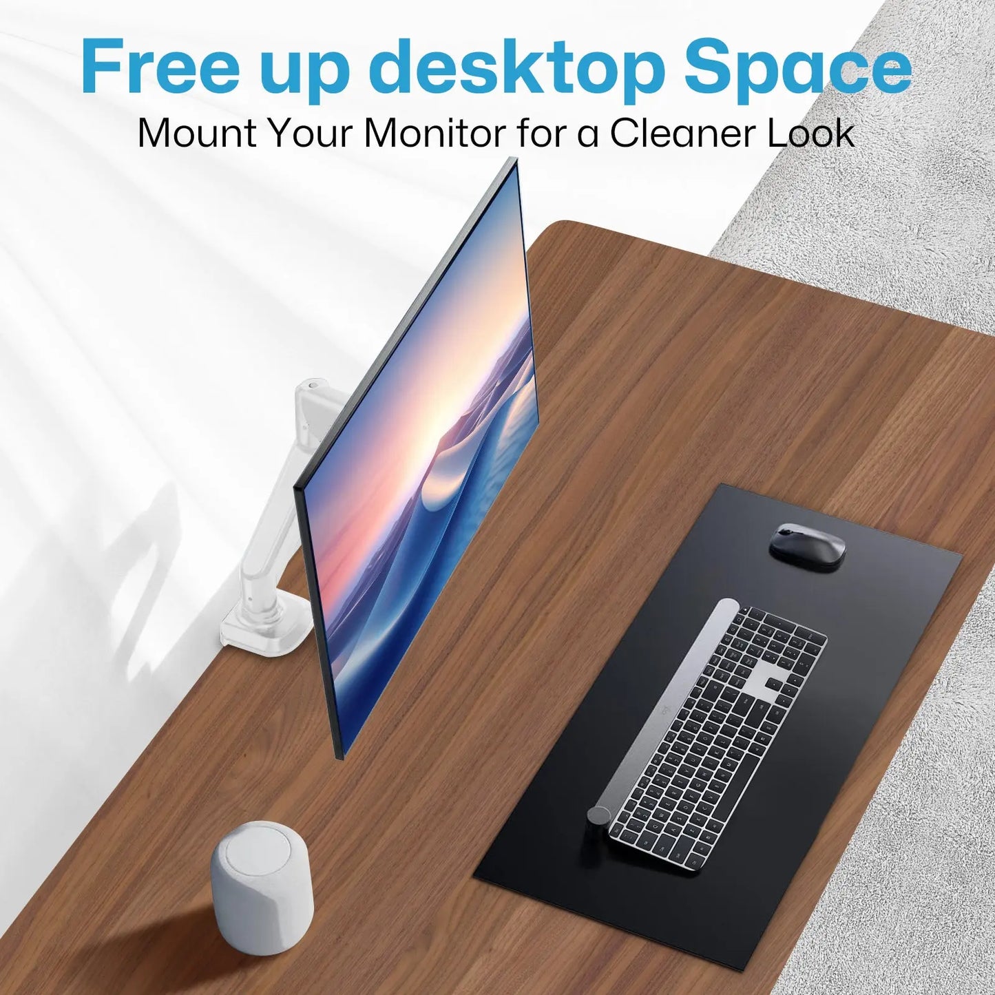 The image shows a clean and organized desktop setup. A monitor is mounted on a white arm attached to the left side of the wooden desk, freeing up space on the desktop. The monitor displays a serene image of waves. On the desk, there is a black mat with a keyboard and a mouse placed on it. A small white speaker or smart device is also present on the desk. The text at the top of the image reads: "Free up desktop Space. Mount Your Monitor for a Cleaner Look."