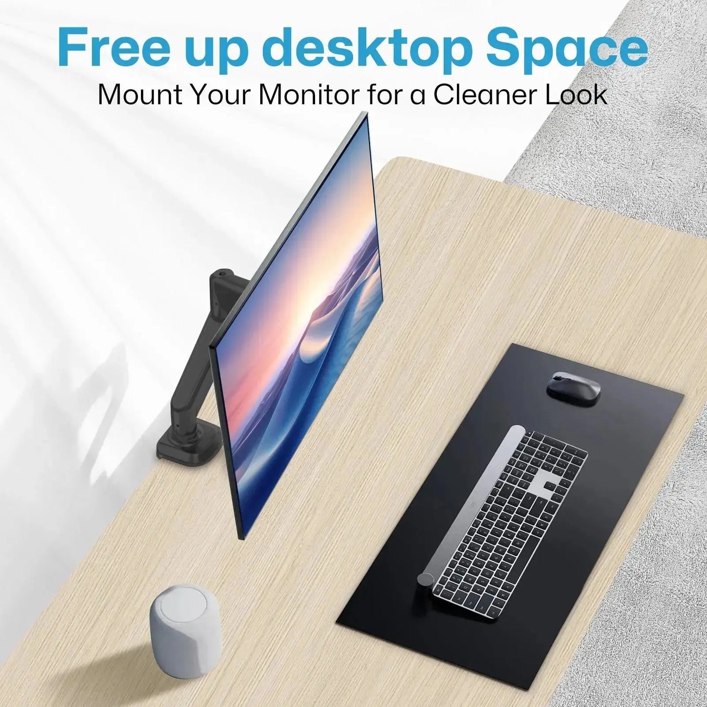 The image shows a clean and organized desktop setup. A monitor is mounted on a white arm attached to the left side of the wooden desk, freeing up space on the desktop. The monitor displays a serene image of waves. On the desk, there is a black mat with a keyboard and a mouse placed on it. A small white speaker or smart device is also present on the desk. The text at the top of the image reads: "Free up desktop Space. Mount Your Monitor for a Cleaner Look."
