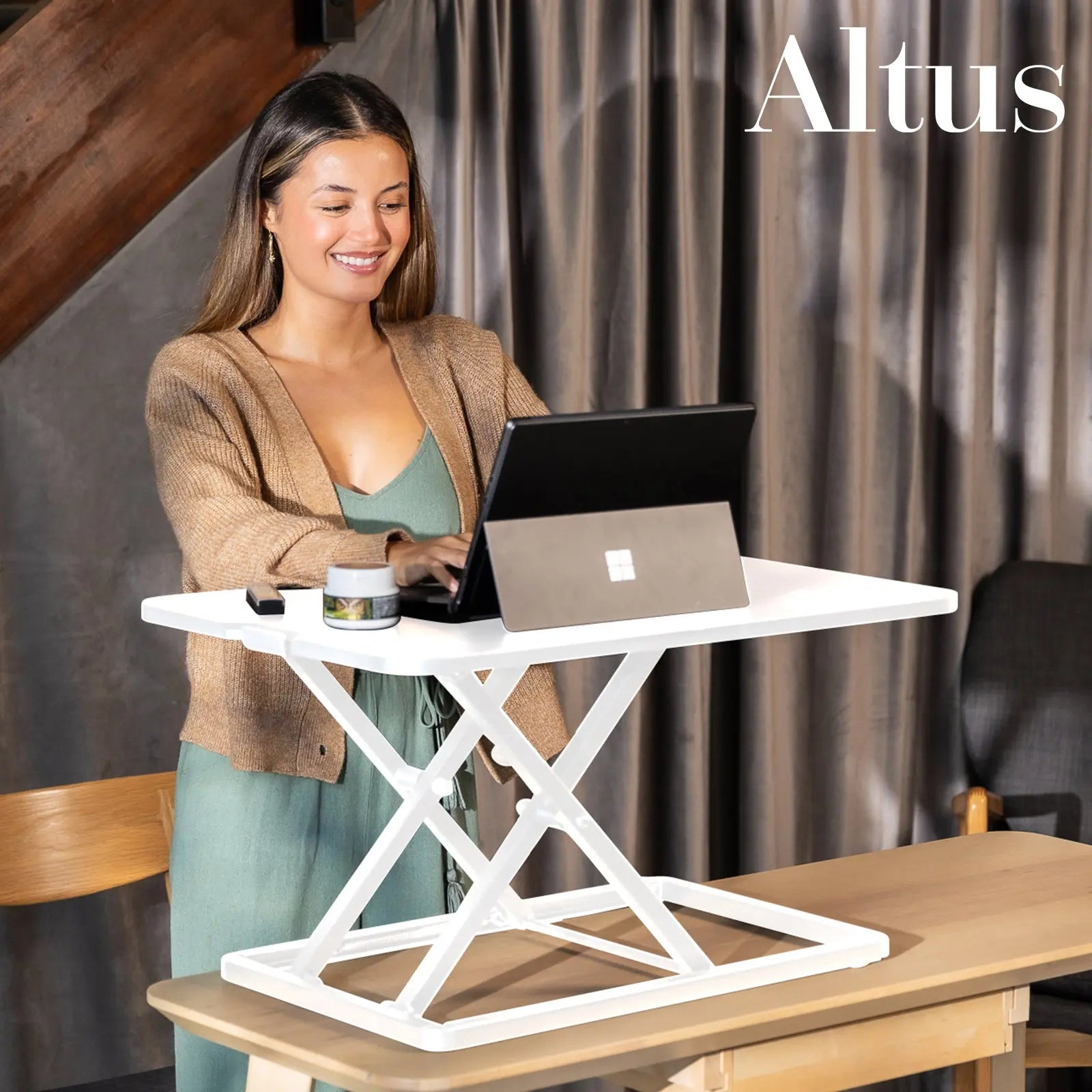 A young woman in a casual outfit stands smiling while working on a laptop placed on a white standing desk in a cozy, modern indoor setting.