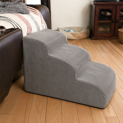 A gray three-step pet ramp with a textured, ribbed fabric surface, shown in a home environment next to a brown leather sofa on a wooden floor.
