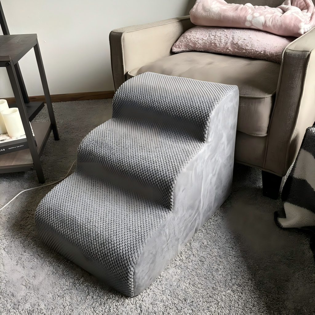A three-step gray pet ramp with a textured, waffle-like surface, positioned next to a beige armchair in a living room setting.