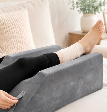 A person reclining on a gray wedge-shaped leg rest cushion on a bed, with one bare foot resting on the cushion. The person is wearing black leggings and a white ankle wrap, with a neutral bedroom background including a pillow and a side table.
