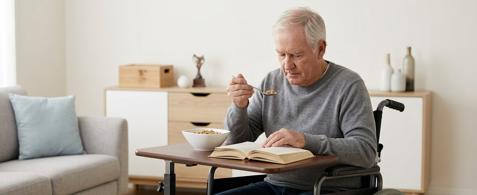 An older man with gray hair sits in a wheelchair at a small table in a bright room, eating from a bowl while reading an open book. A sofa and cabinet are visible in the background.