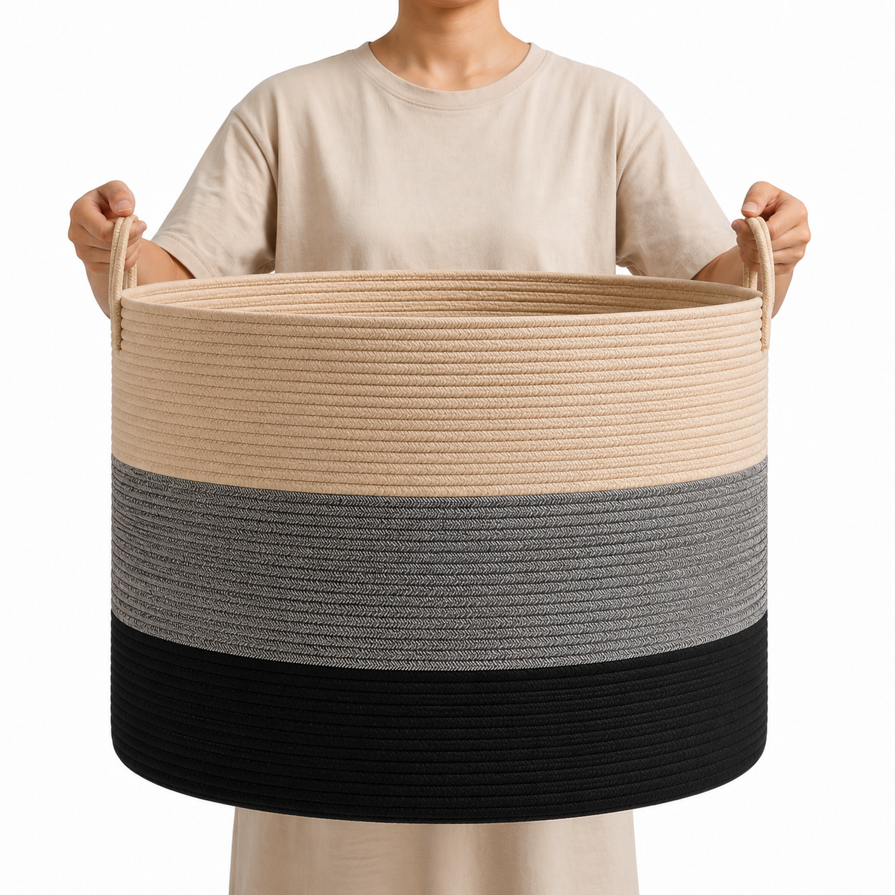 A person holding a large rope storage basket with three horizontal color bands: beige on top, gray in the middle, and black on the bottom, against a plain white background.