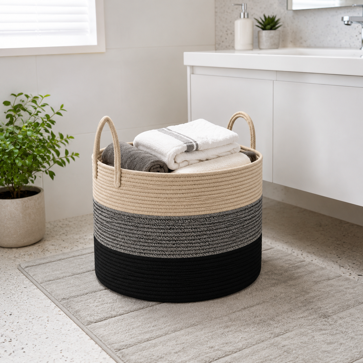A round woven storage basket with two handles sits on a bathroom rug in a bright bathroom. The basket has a three-tone striped design in beige, gray, and black, and is filled with folded white and gray towels.