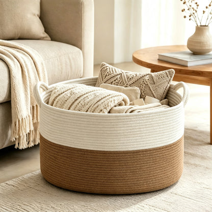 A round woven storage basket with a two-tone design, featuring a cream upper band and a natural brown rope lower half. The basket is filled with neutral-toned knit blankets and pillows, set in a bright, modern living room next to a sofa and a wooden side table.