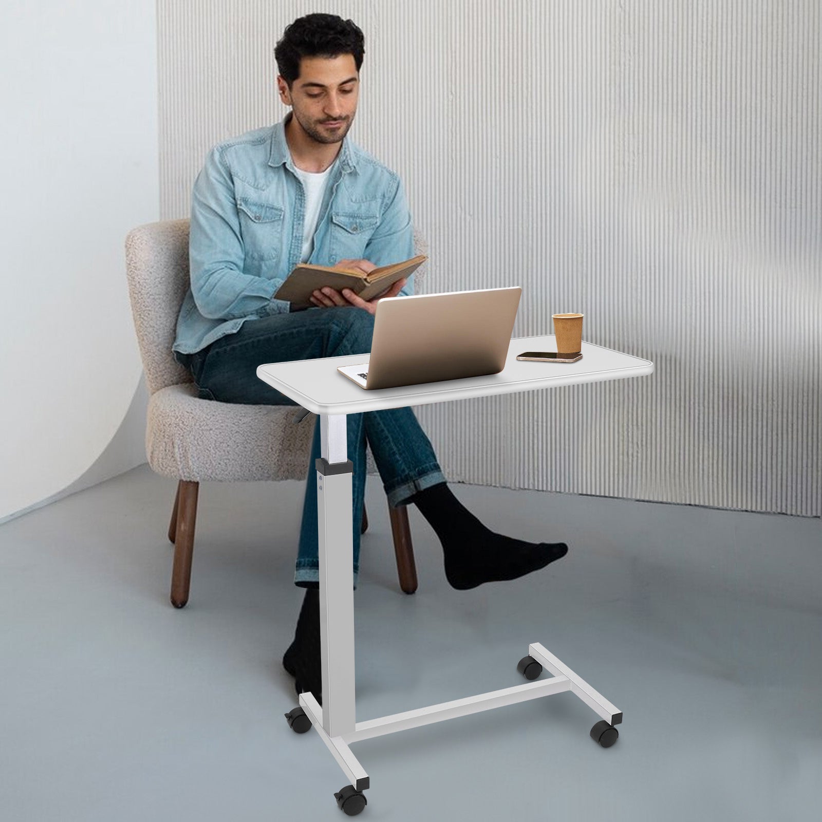 Man sitting at a white adjustable desk with a laptop and tablet, holding a book.