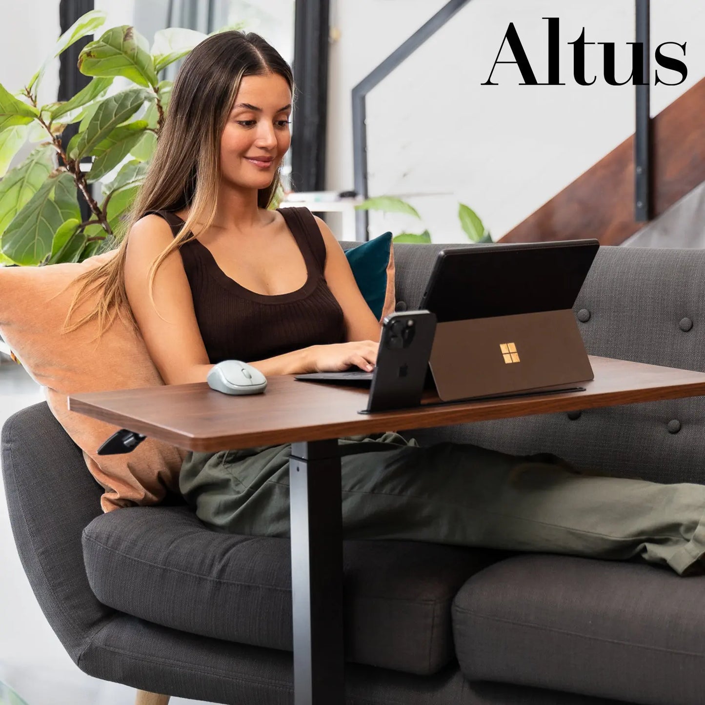 A young woman working on a laptop at a mobile standing desk positioned beside a comfortable sofa in a modern home office.