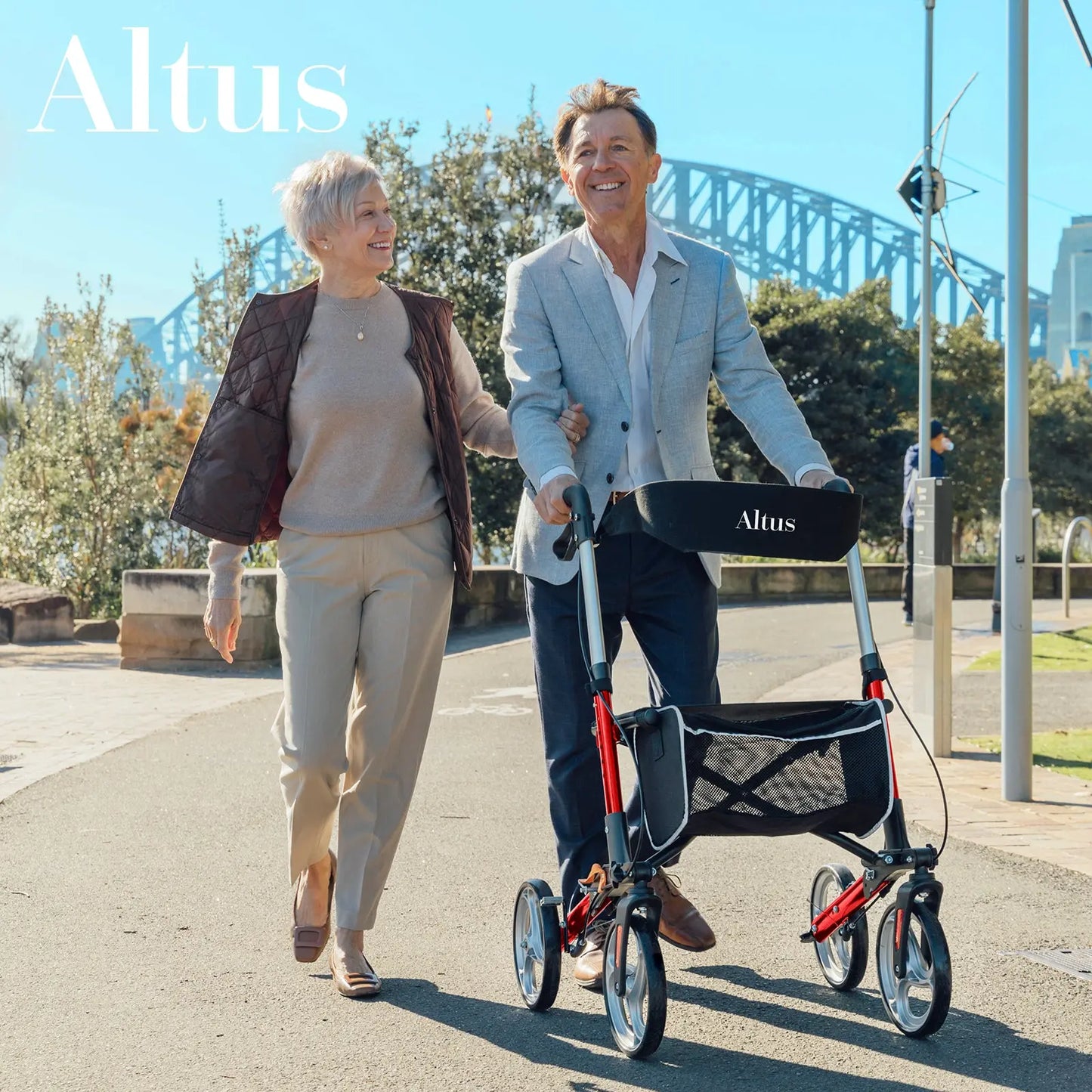 An elderly couple is walking outdoors with the man using the Altus Euro Style Rollator. The couple is smiling and enjoying a sunny day, demonstrating the rollator's role in promoting mobility and independence. The rollator is red with a black seat and storage bag, offering support and convenience. The scenic background with a bridge adds to the appeal, showing the rollator's suitability for various environments.