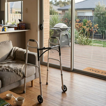 A silver metal rolling walker with black handles and wheels is positioned on a wooden floor in a home interior, with a sofa, kitchen area, sliding glass doors, and outdoor deck visible.