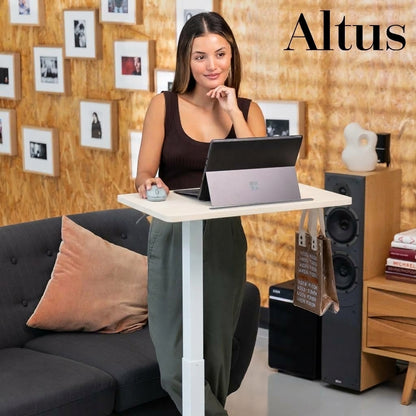 A woman stands using an Altus standing desk in a modern living room setting. The white T-shaped standing desk supports a laptop, with a couch, speakers, and a wood-paneled wall in the background.
