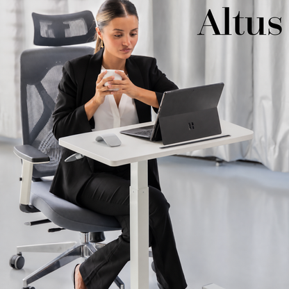 A woman in a black blazer sits in a gray mesh office chair at a white AltUS standing desk, using a laptop and holding a mug in a bright office setting.