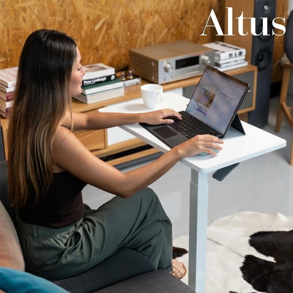 A woman sits on a couch using a tablet on a white Altus standing desk in a modern home office setting with wood paneling.