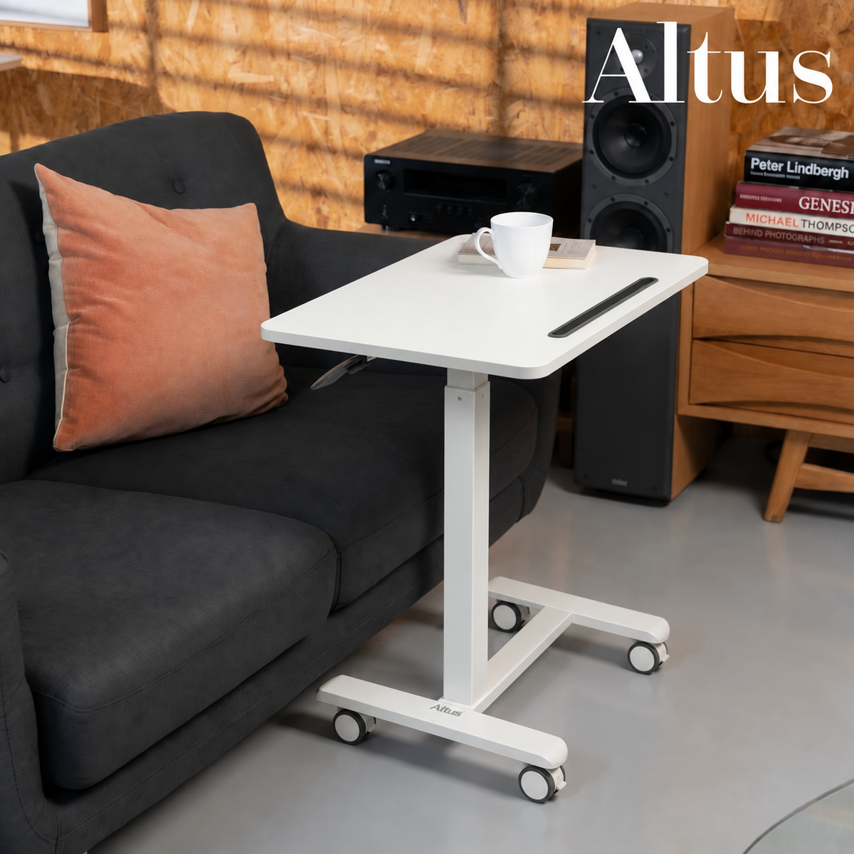 A white adjustable height standing desk with wheels, styled next to a dark gray sofa in a modern living room setting. The desk features a white top with a black pen holder and a white mug on top.
