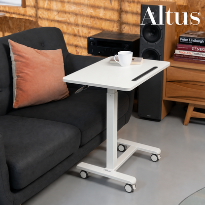 A white adjustable height standing desk with wheels, styled next to a dark gray sofa in a modern living room setting. The desk features a white top with a black pen holder and a white mug on top.