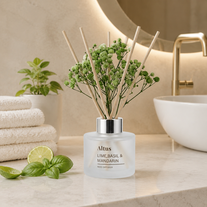 Altus reed diffuser bottle with green baby's breath and natural reed sticks, set on a bathroom counter with a sink, folded towels, basil, and lime slices.