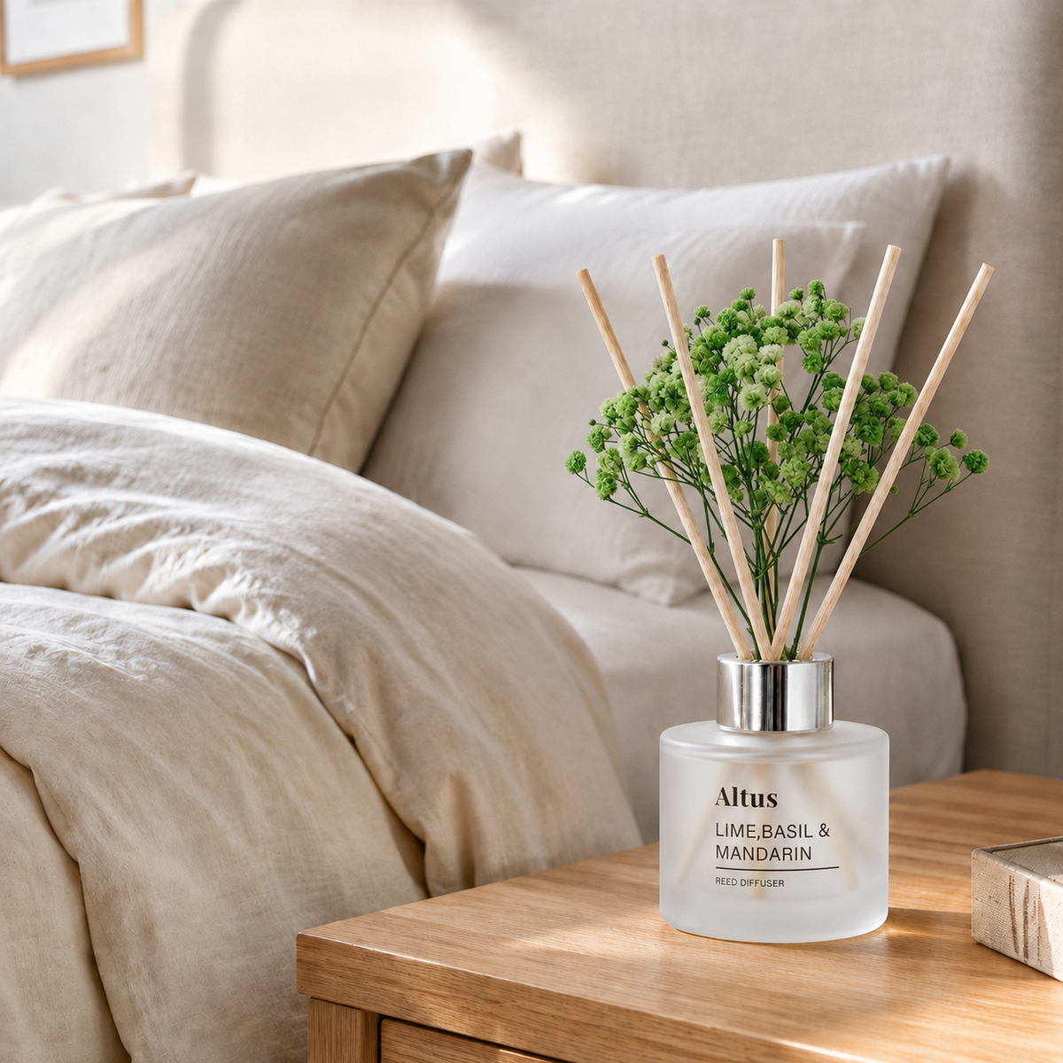 Altus Lime-Basil & Mandarin reed diffuser bottle with green reeds and dried flowers, styled on a wooden bedside table next to a bed with beige bedding.