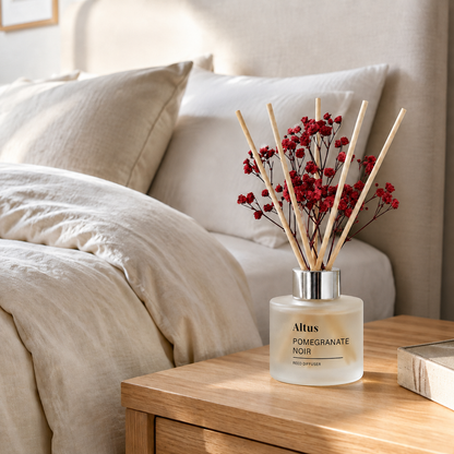 Altus Pomegranate Noir reed diffuser bottle with natural reeds and dried red berries, styled on a wooden bedside table next to a bed with beige bedding.