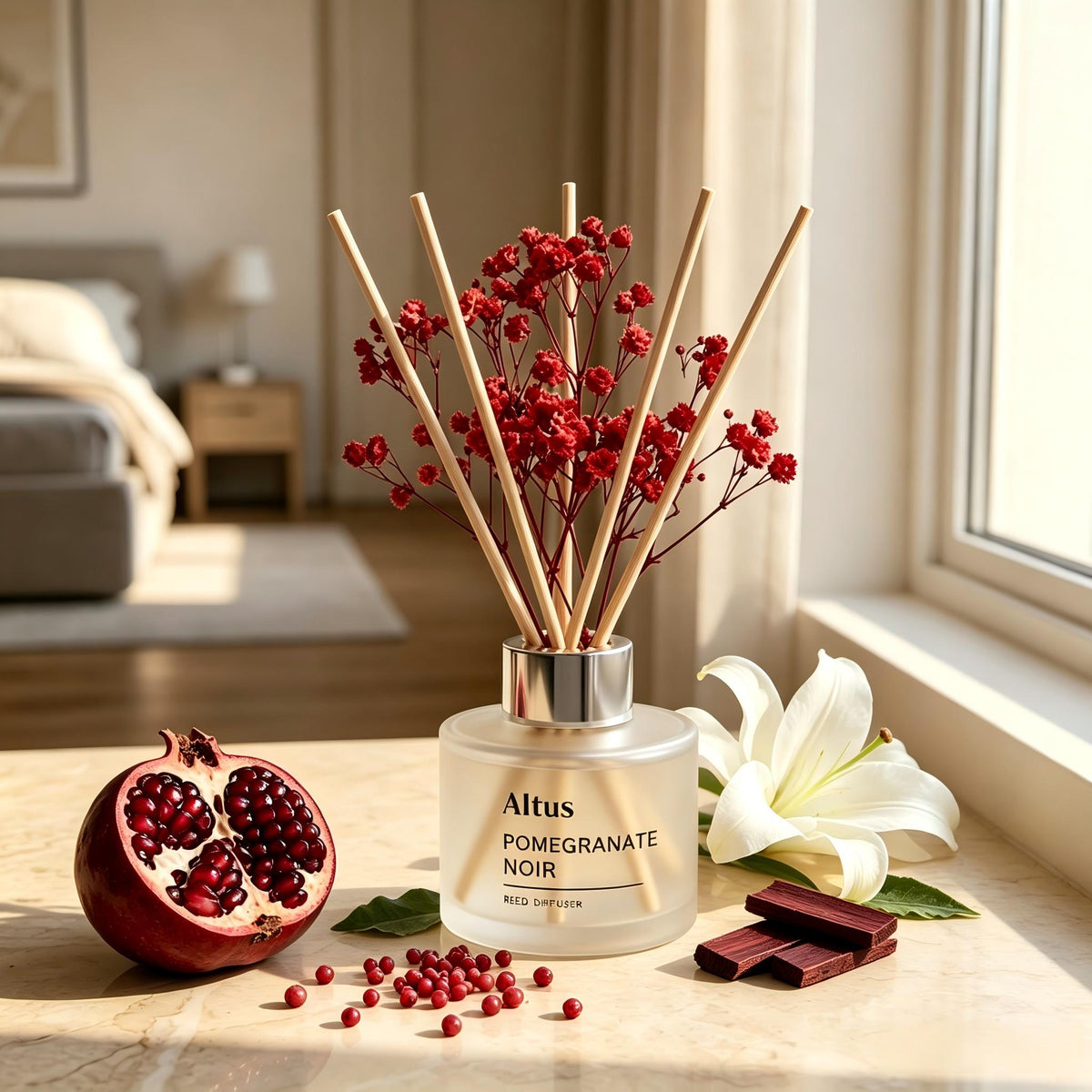 Altus Pomegranate Noir reed diffuser bottle with wooden reeds and red berries, styled on a table with a pomegranate half, loose seeds, chocolate pieces, and a white lily flower in a bright bedroom setting.