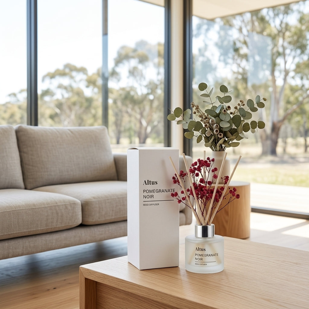 Altus Pomegranate Noir reed diffuser bottle with reeds and a matching white product box, styled on a wooden table in a modern living room with a sofa and large windows.