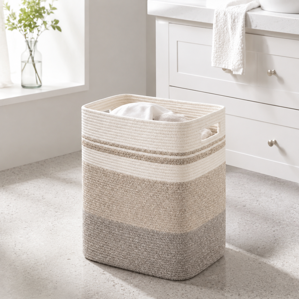A rectangular woven rope laundry hamper basket in horizontal cream, beige, and taupe-gray stripes, filled with white towels and placed on a light gray speckled floor in a bright bathroom next to a white vanity.