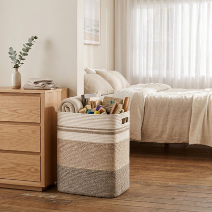 A woven rectangular laundry hamper basket in beige and cream stripes sits on a wooden floor in a softly lit bedroom, filled with rolled towels and stationery.