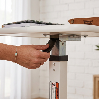 A close-up of a person's hand adjusting the black lever on a white ANKURA height-adjustable table leg, with a white tabletop and a notebook in the background.