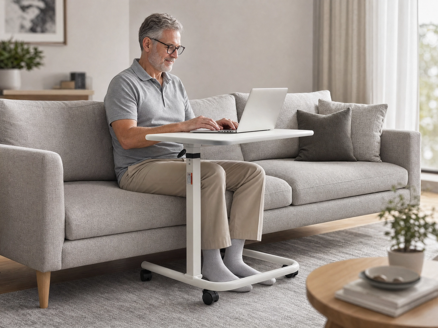 A man sits on a light gray sofa in a bright living room, using a white lap desk with a built-in laptop. The lap desk has a white top and base with black caster wheels, and the man is wearing a gray polo shirt and beige pants.